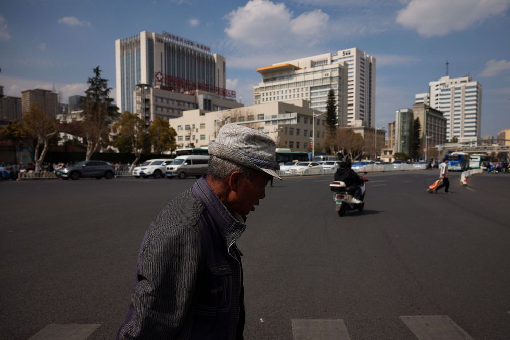 A person wearing a hat walks past modern office buildings in an urban setting on a sunny day.