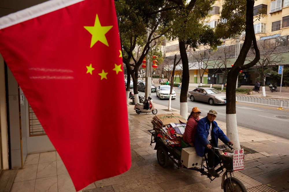 Chinese national flag waves prominently on a city street as people rest on benches in the shade of trees.