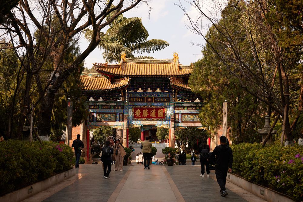 Visitors walk along a path leading to a traditional Chinese temple gate with ornate architecture and golden roof tiles.