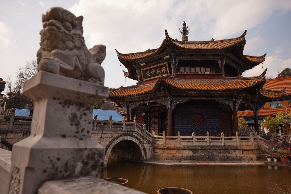 Traditional Chinese temple with ornate curved roofs and stone lion guardian statue sits beside a decorative bridge over water.