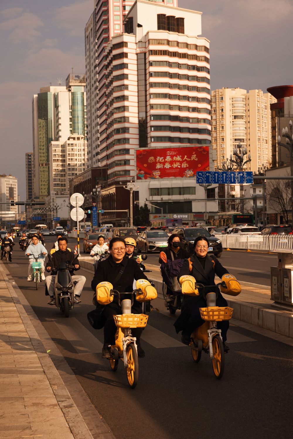 Commuters ride yellow delivery scooters along a busy street in China with modern high-rise buildings in the background.