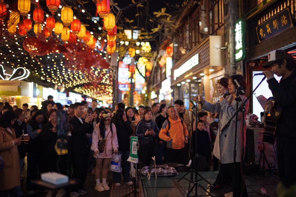 Crowded nighttime street in Chinatown decorated with colorful lanterns and string lights during a festive celebration.
