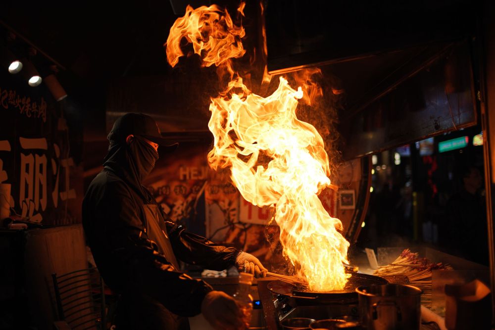 A chef creates dramatic flames while cooking at a street food stall at night.