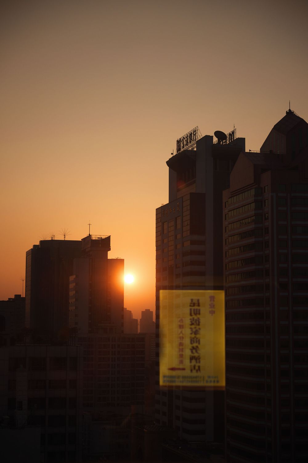 Silhouette of city buildings against an orange sunset sky with a yellow advertisement banner visible between towers.