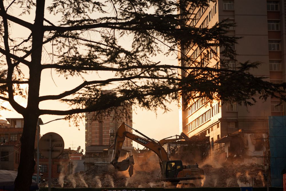 Silhouetted industrial machinery and construction cranes against a glowing sunset backdrop with city buildings.
