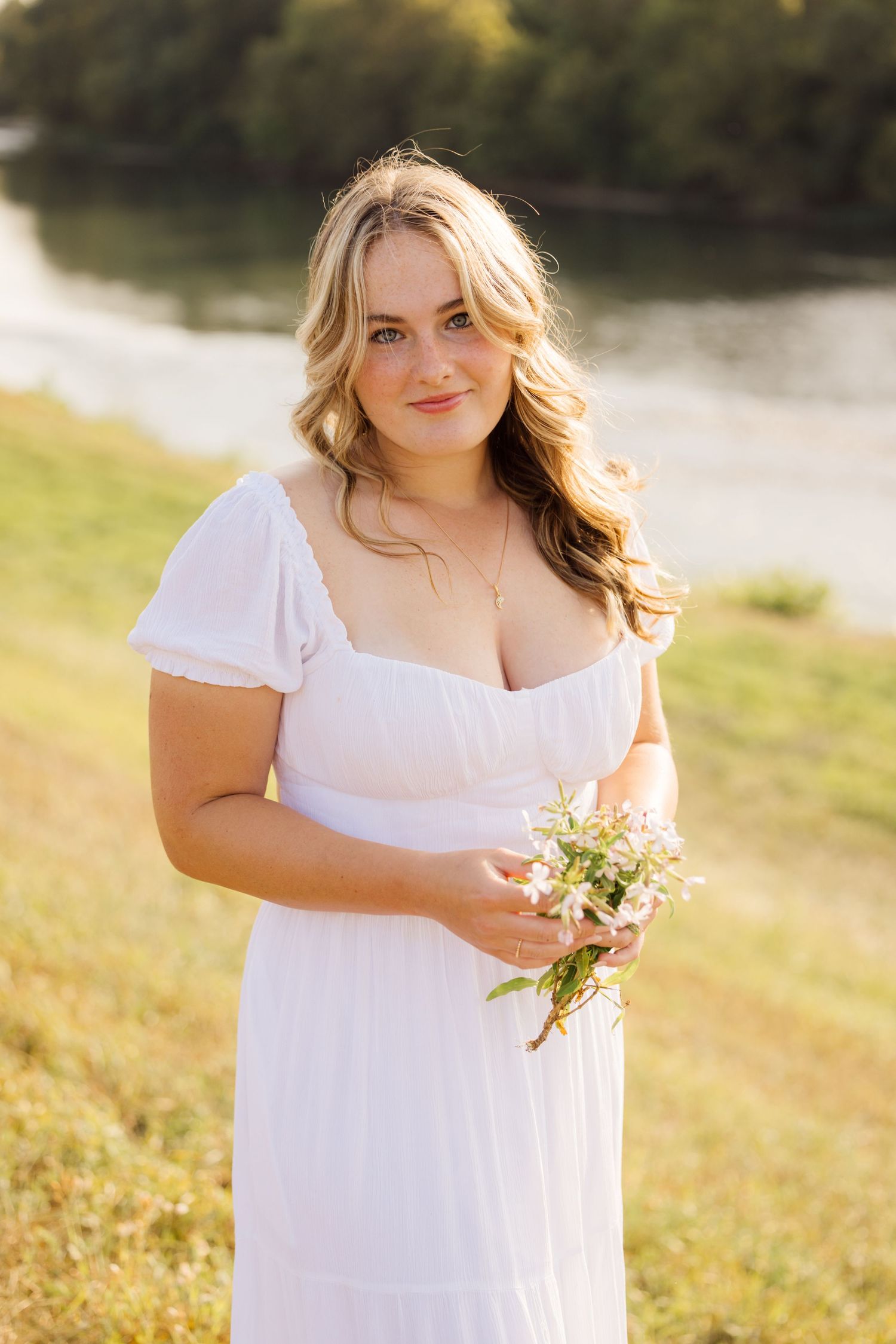A person in a flowing white dress holds wildflowers while standing in a sunlit meadow near a lake.