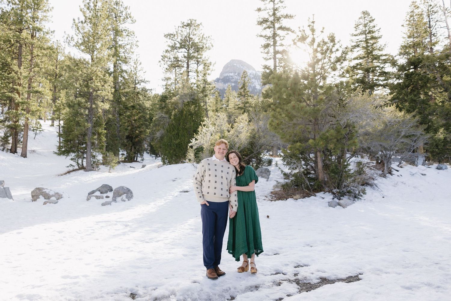 Two people stand together on a snowy mountain path with evergreen trees and a mountain peak visible in the background.