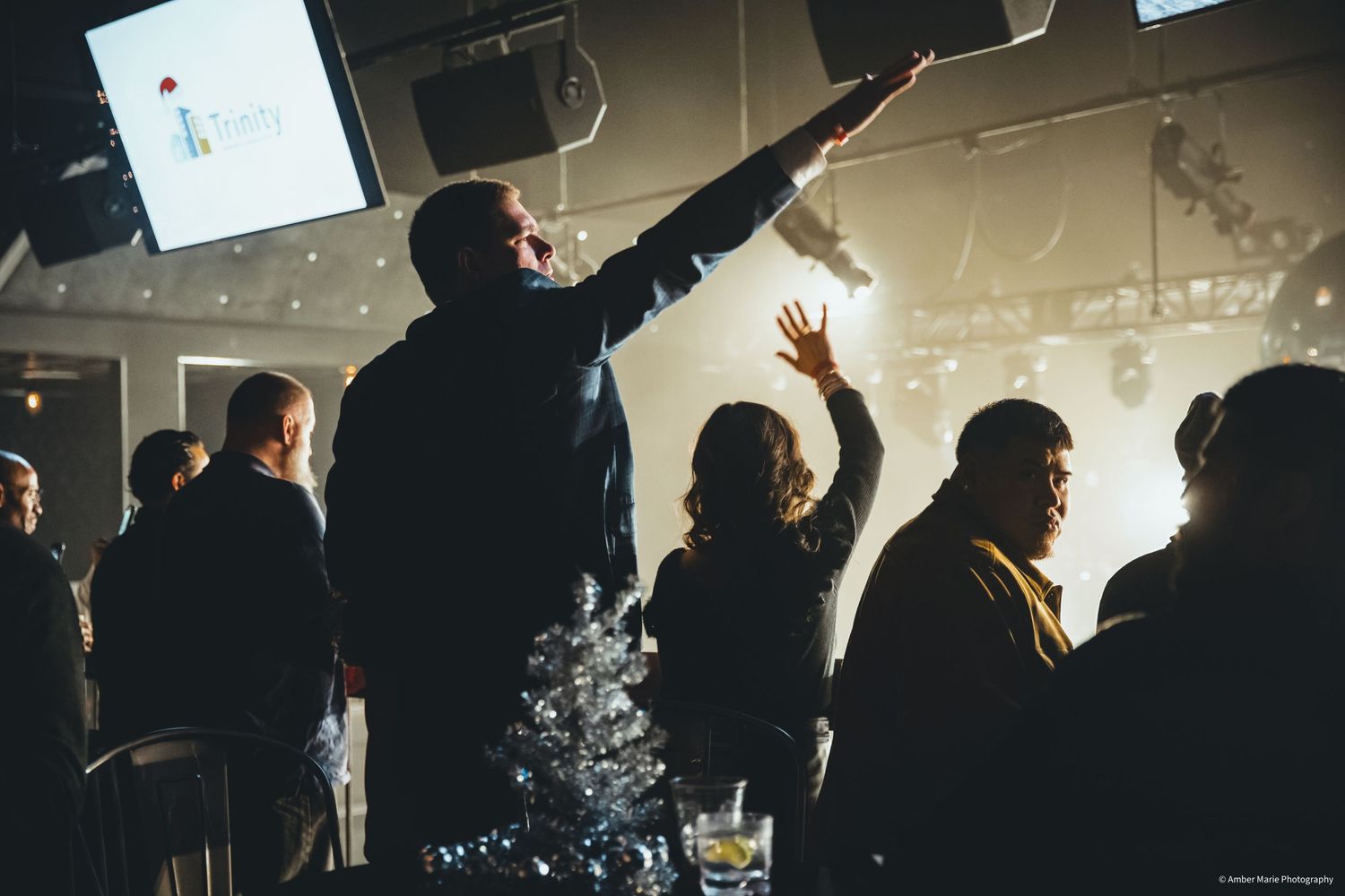 Silhouetted crowd raising hands at a dimly lit indoor event with monitors visible overhead.