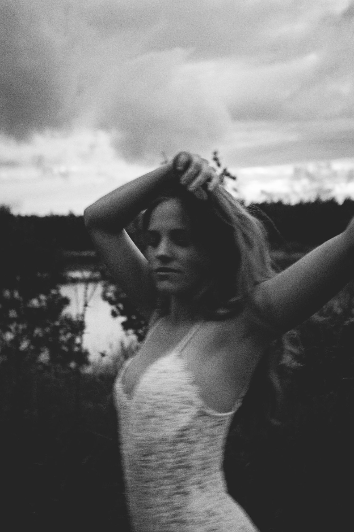 Black and white portrait of someone with wind-swept hair against a cloudy sky and water backdrop.