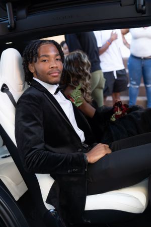 Person in black tuxedo sitting in white leather car seat at formal event.