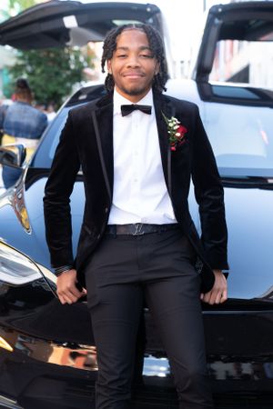 Person in velvet tuxedo and bow tie posing in front of black Tesla car.