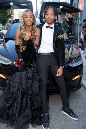 Formal couple dressed in black evening wear standing beside a Tesla car.