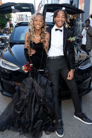 Elegant couple poses by luxury car trunk at formal event in black dress and tuxedo with floral boutonniere.