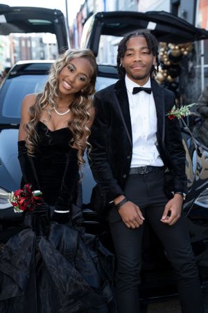 Couple smiles for photo at formal event wearing black velvet gown and suit in front of car.
