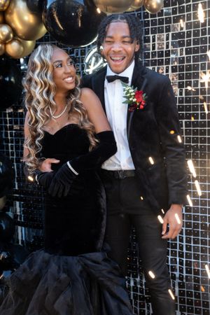 Glamorous prom couple in black formal wear against sparkly backdrop with balloons.