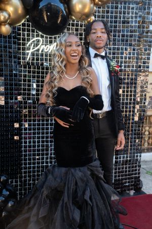 Formal couple in black attire smiling against metallic backdrop with gold balloons.