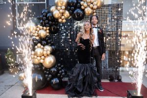 Prom couple posing with sparklers and balloons on red carpet.