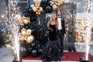 Couple posing in front of black and gold balloon arch with sparkler effects at formal event.