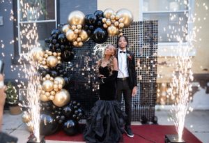 Formal event setup with black and gold balloon arch, sparklers, and glittering backdrop on red carpet.