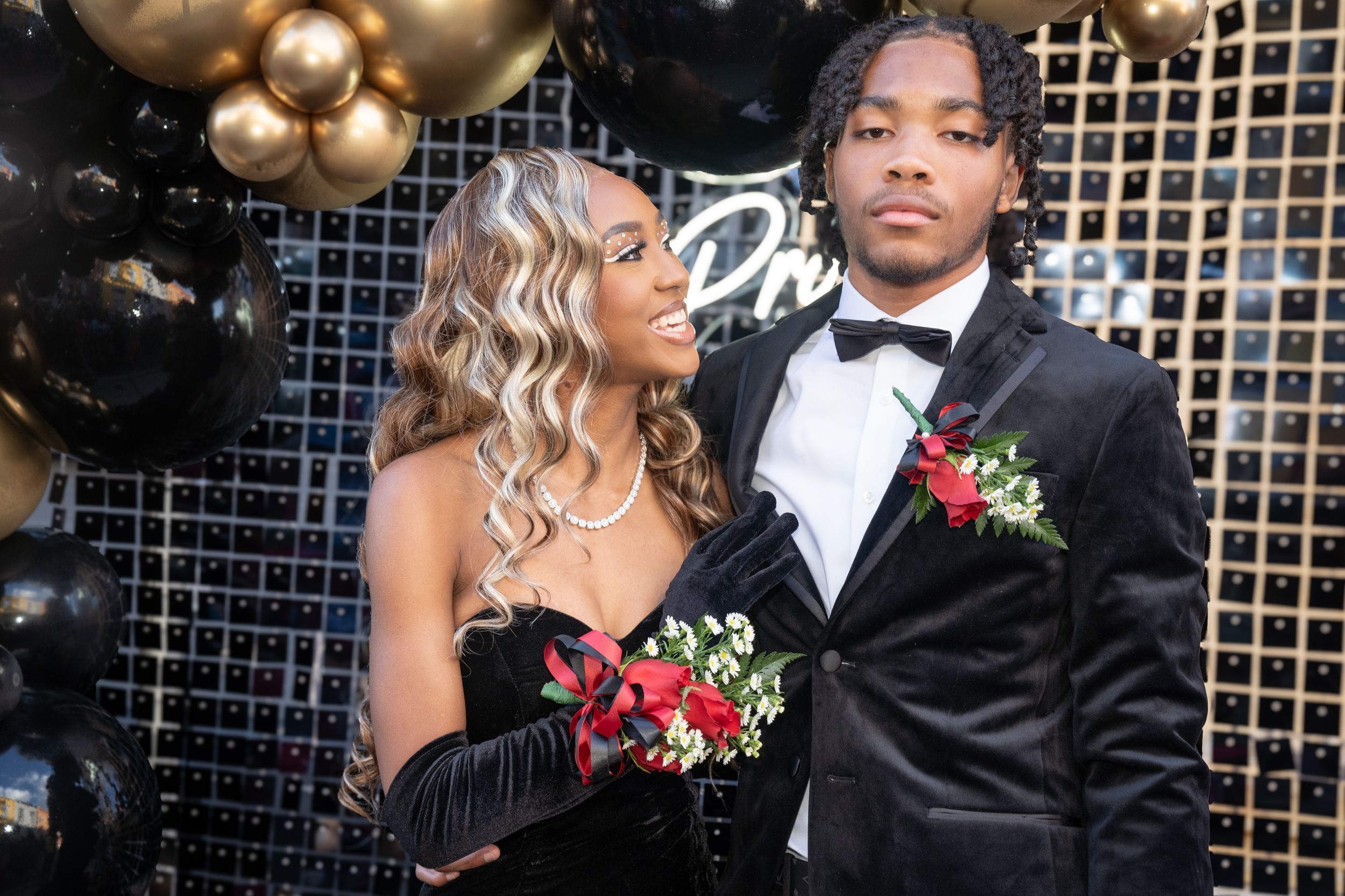 Couple in black formal wear poses at prom with gold balloon backdrop.