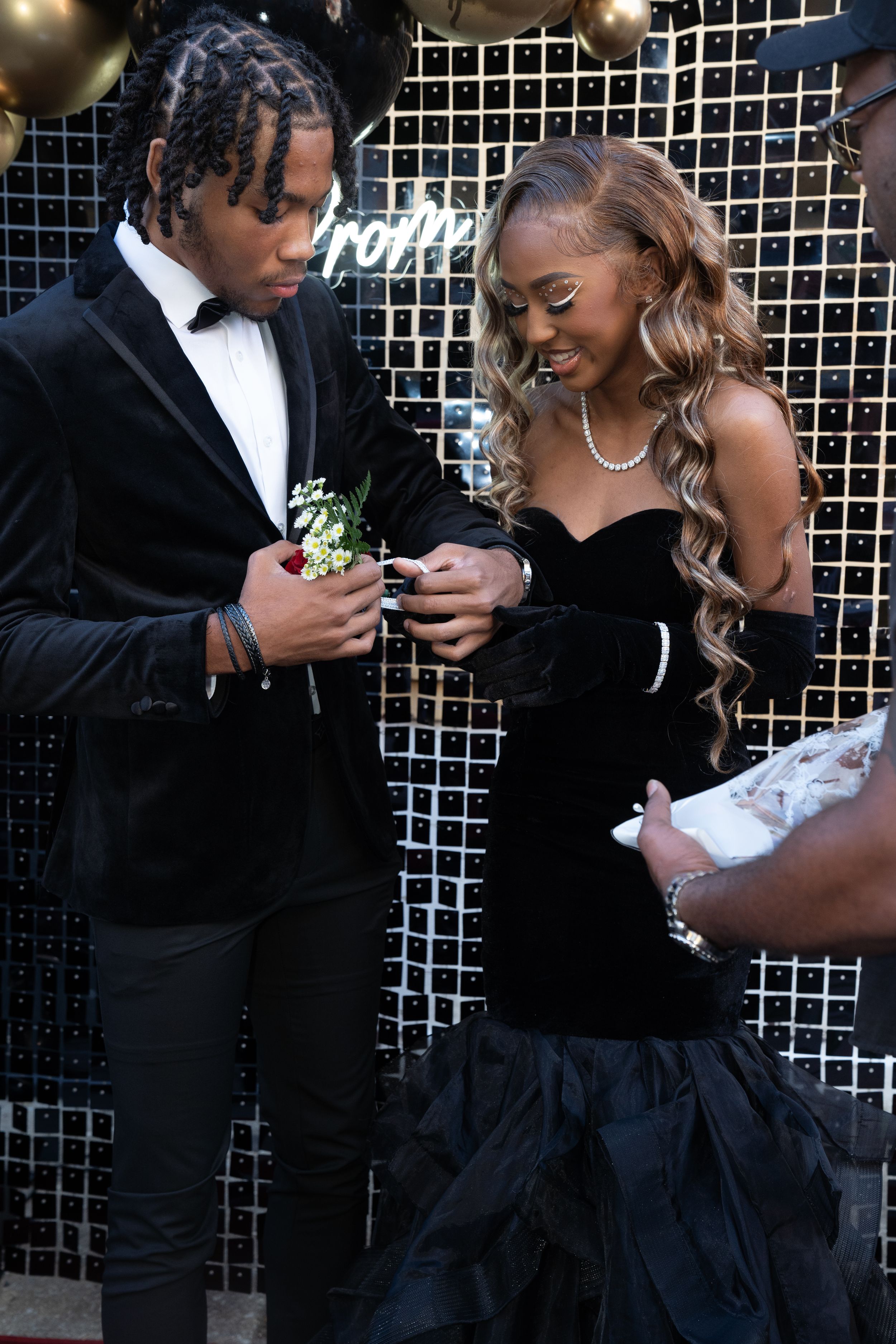 Couple in black formal attire examines corsages at prom event.