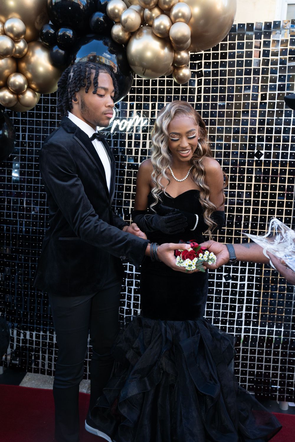 Couple in formal attire receives corsage at prom with balloon decorations.