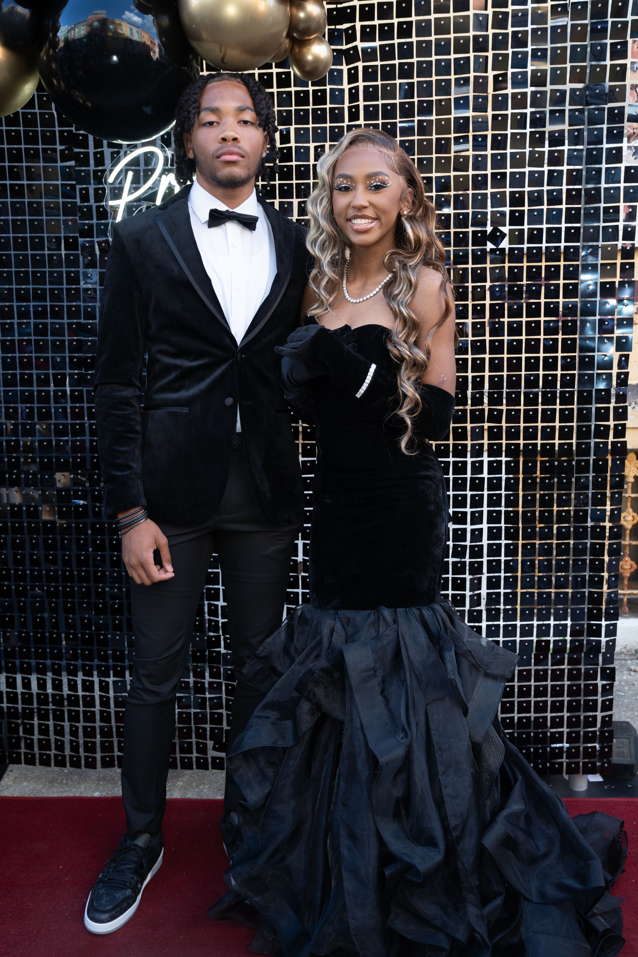 Couple poses on red carpet in matching black formal wear against decorated backdrop.