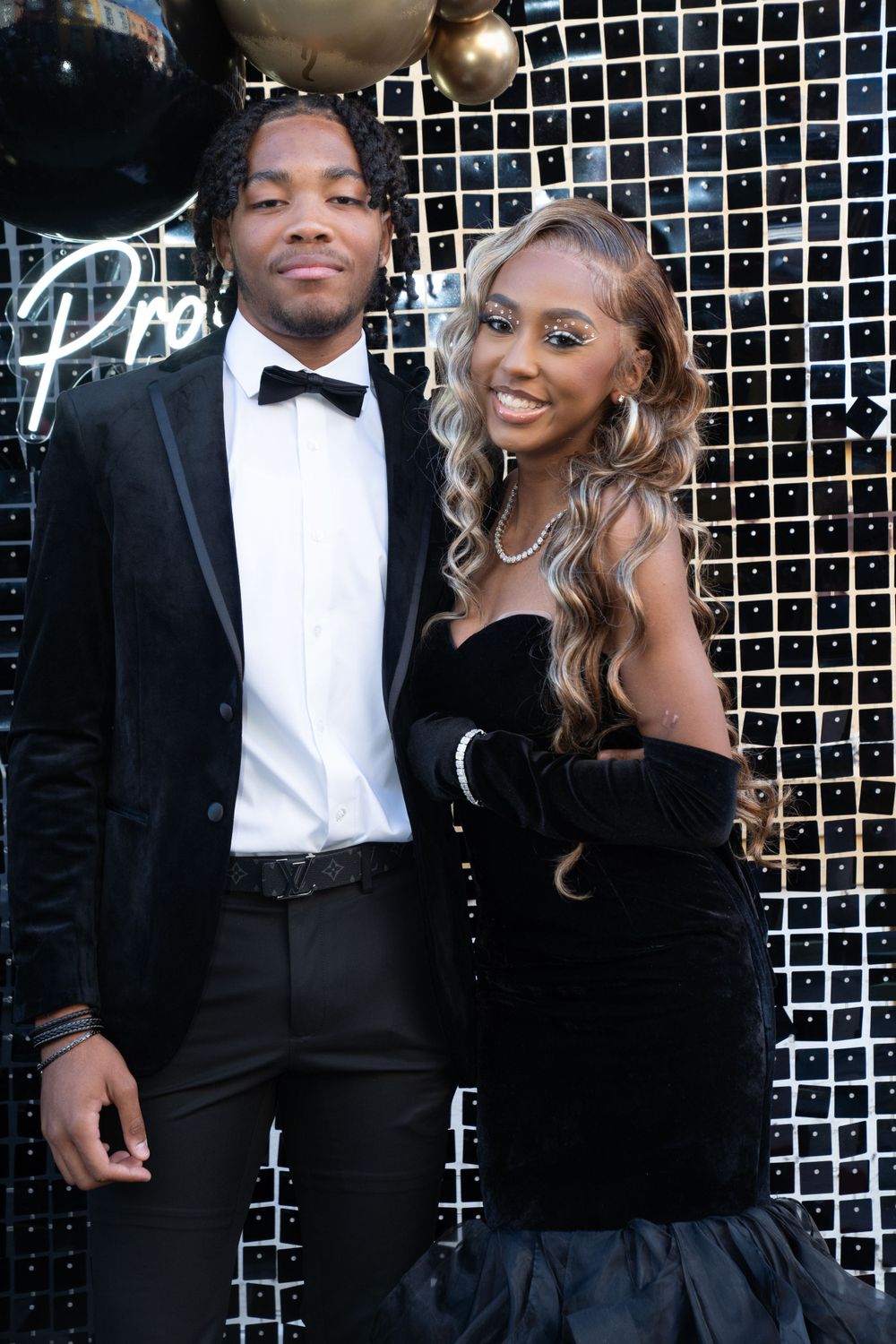 Couple smiles for photo at prom against backdrop with black and gold balloons.