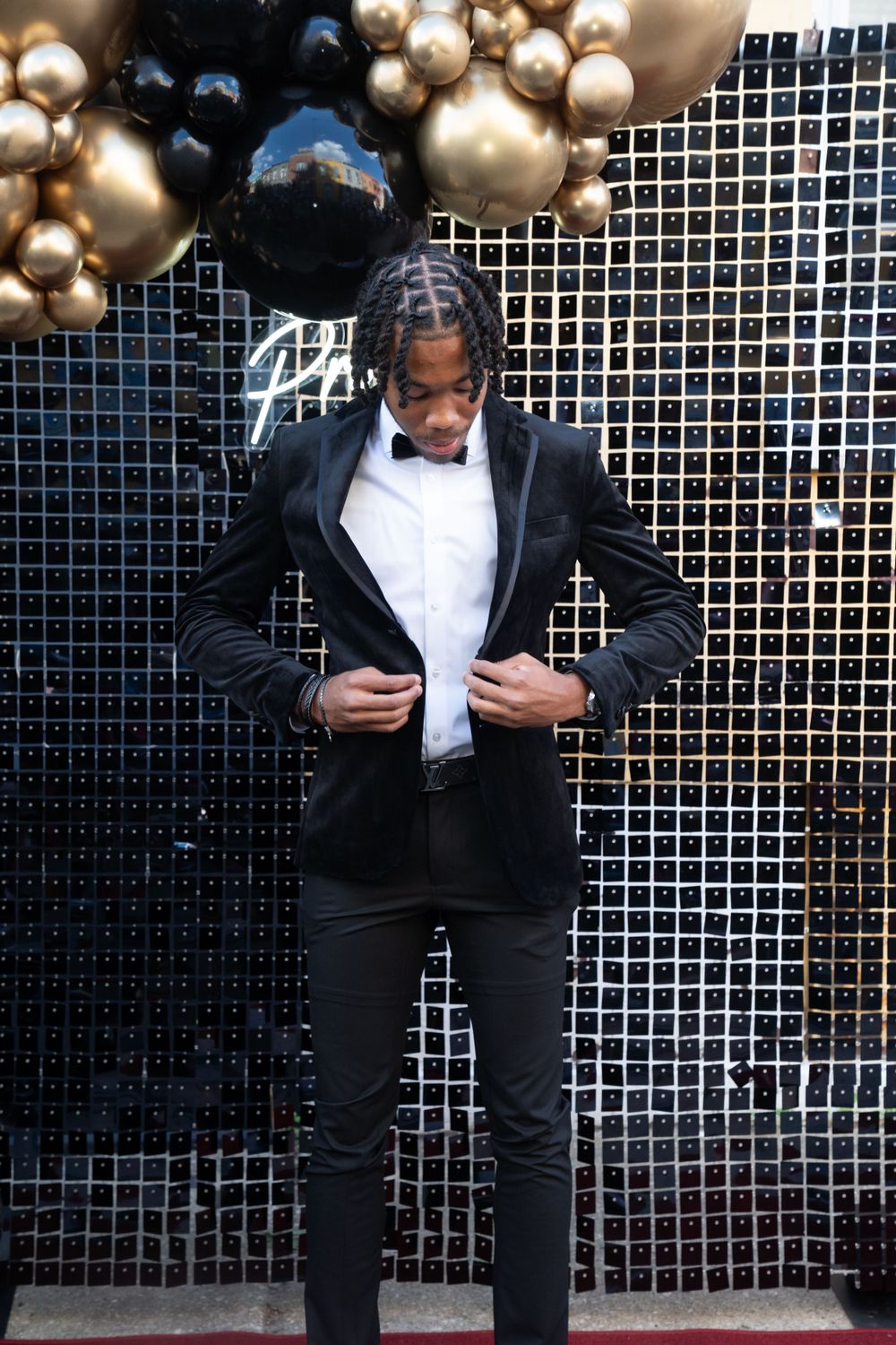 Person in black tuxedo adjusts jacket in front of balloon-decorated wall at formal event.