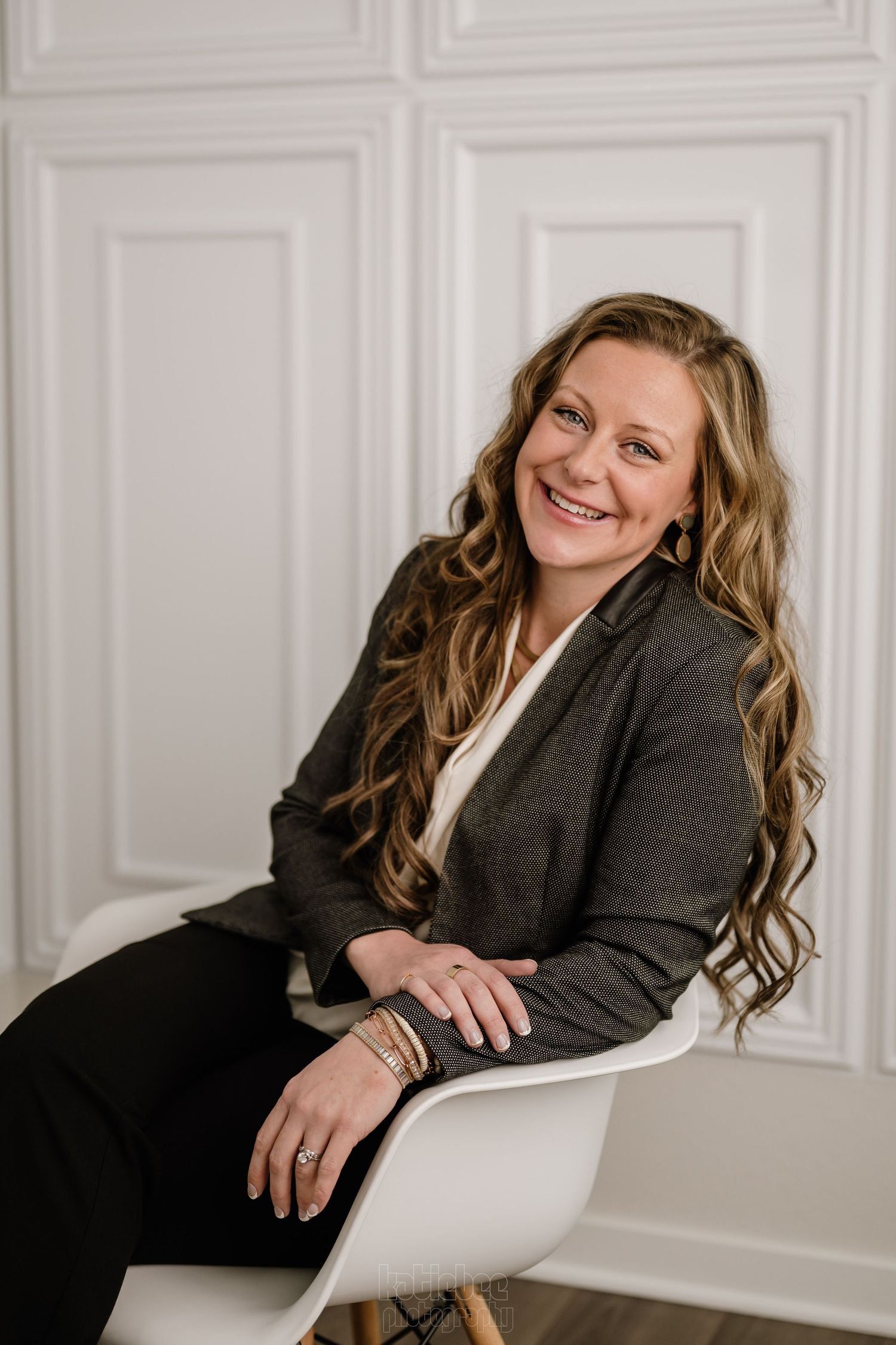 Professional portrait of someone in a dark blazer sitting in a white chair against a white paneled wall with long wavy hair.
