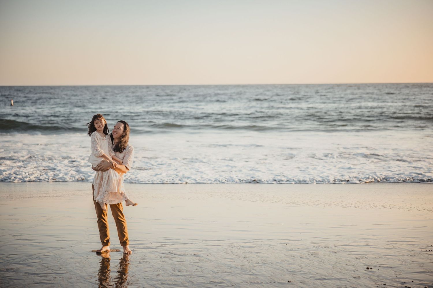 A couple embraces on a serene beach at sunset with ocean waves rolling in behind them.