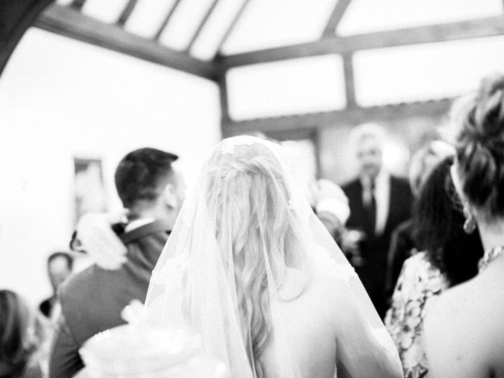 A bride in a white veil is viewed from behind during an indoor wedding ceremony.