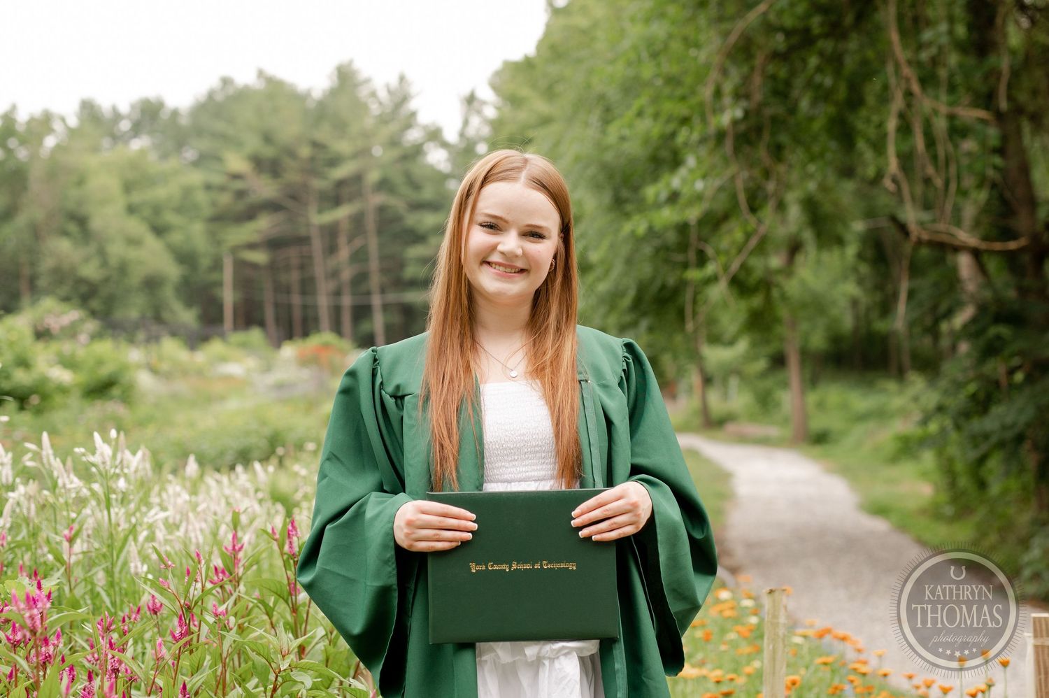 Graduate holding diploma holder poses in green regalia along garden pathway.