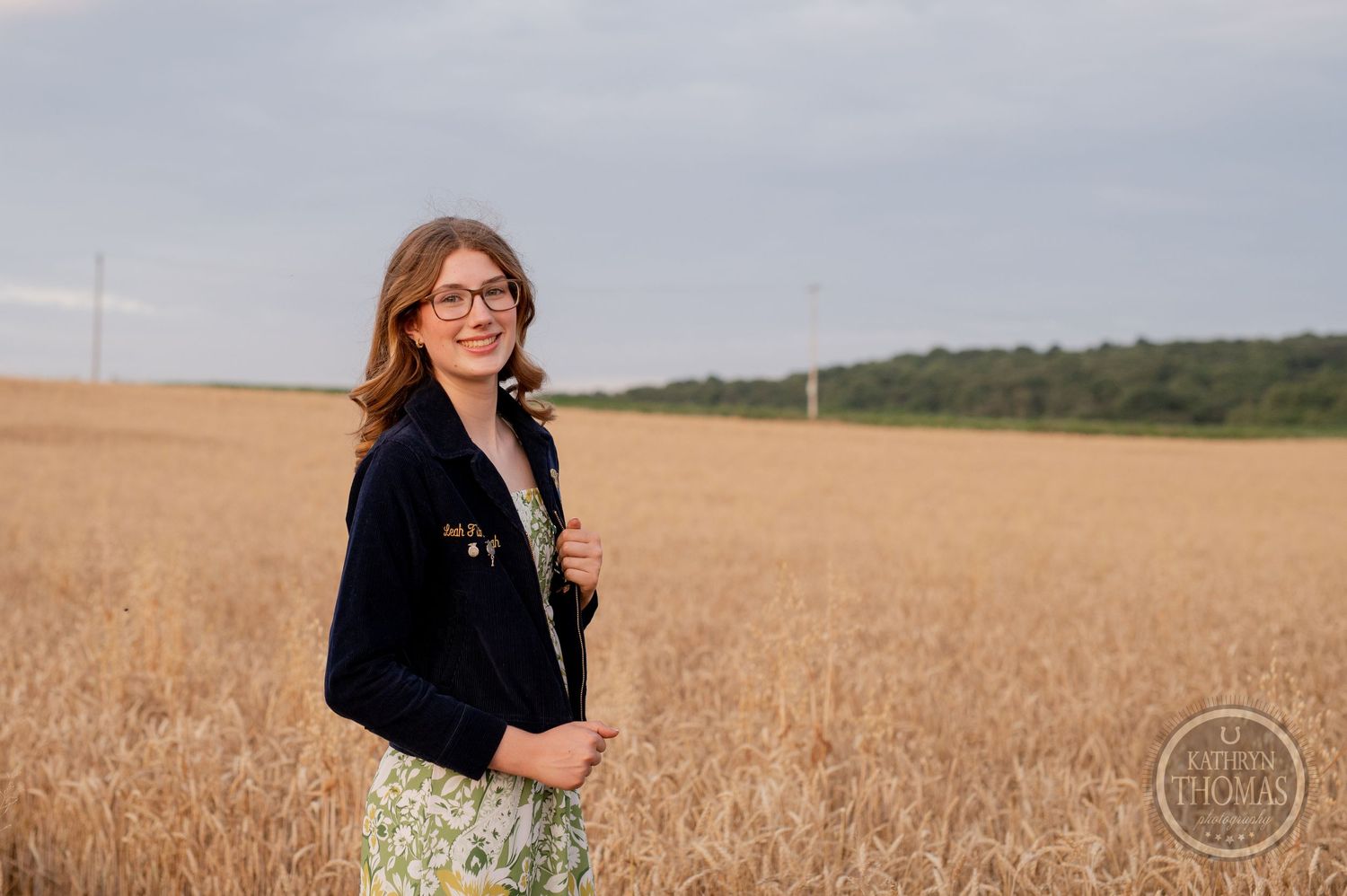 Person wearing FFA jacket and floral dress poses in golden wheat field at dusk.