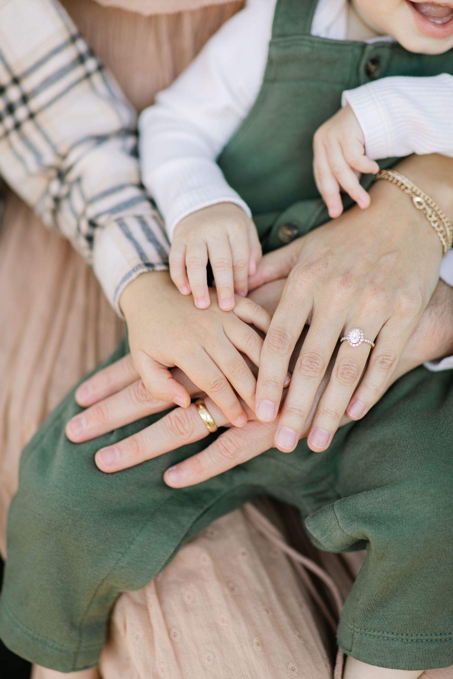 Close-up of hands with a diamond engagement ring showing over a green dress and plaid shirt.Baby and toddler and parents hand pile