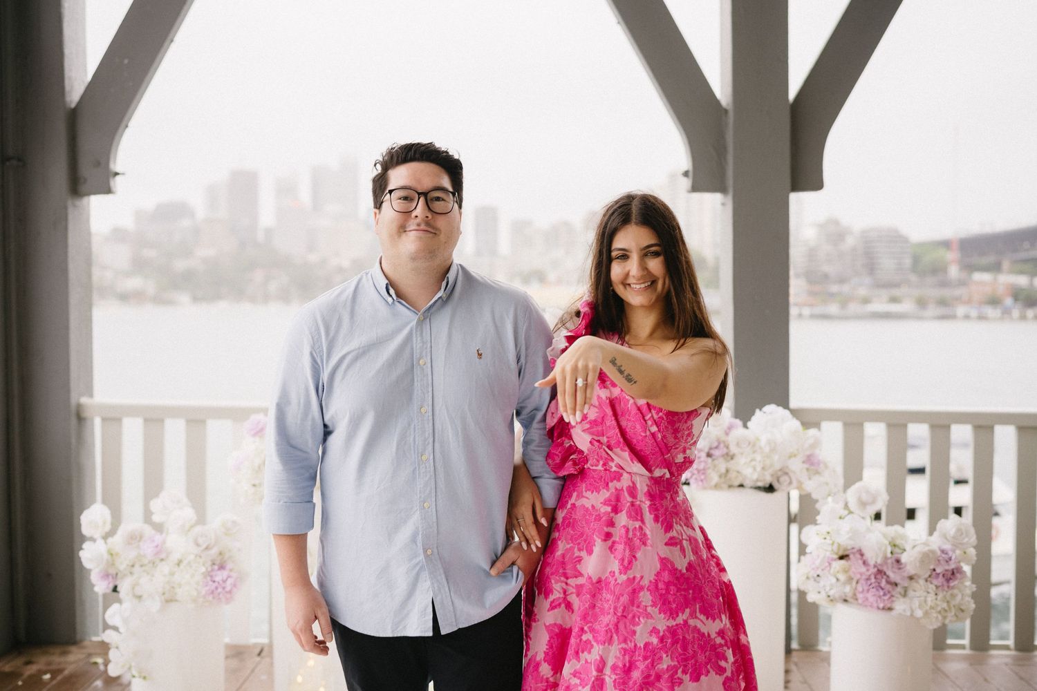 Happy couple posing on waterfront balcony decorated with flowers, woman in pink floral dress.