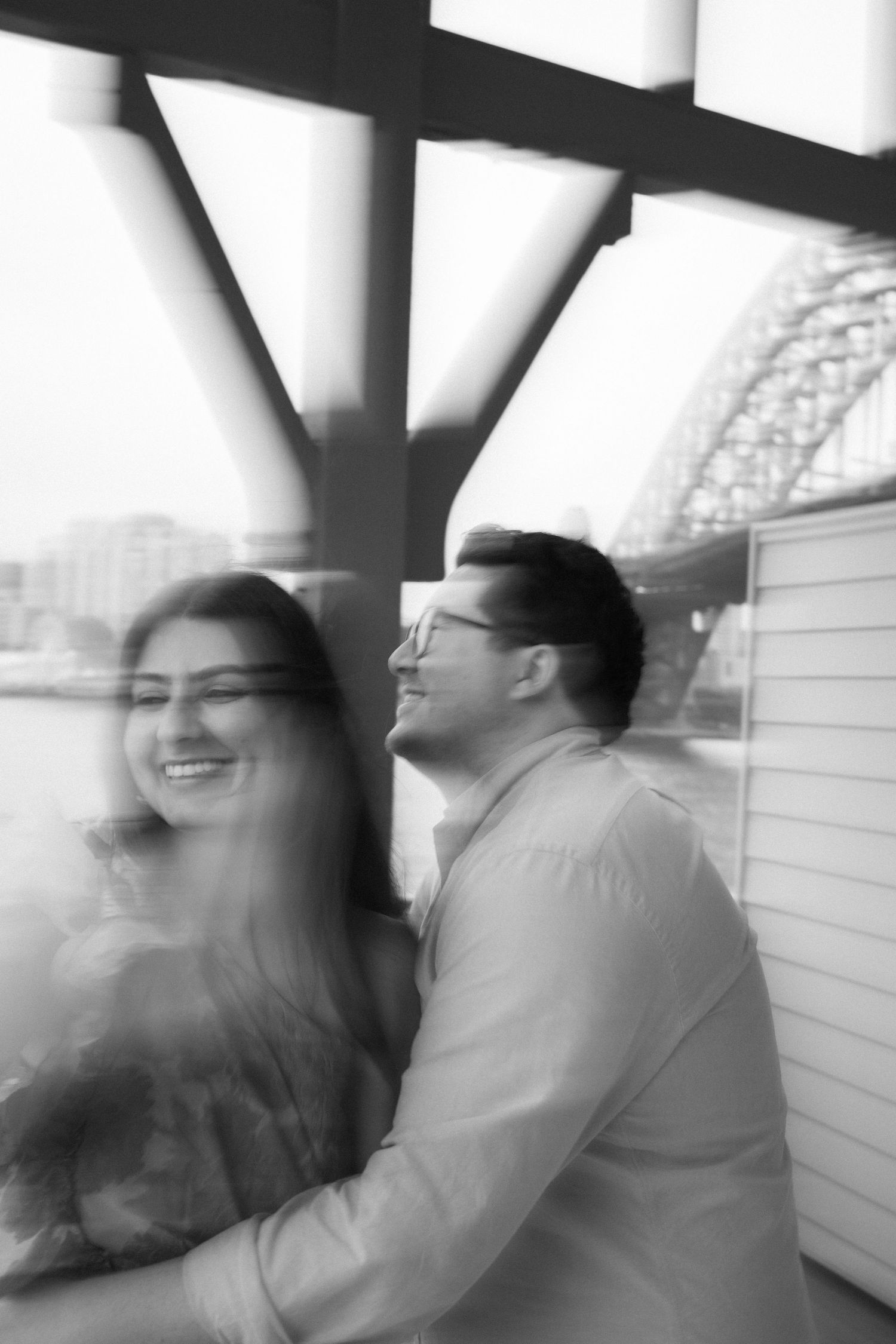 A black and white photo captures a joyful couple sharing a laugh under an architectural structure.