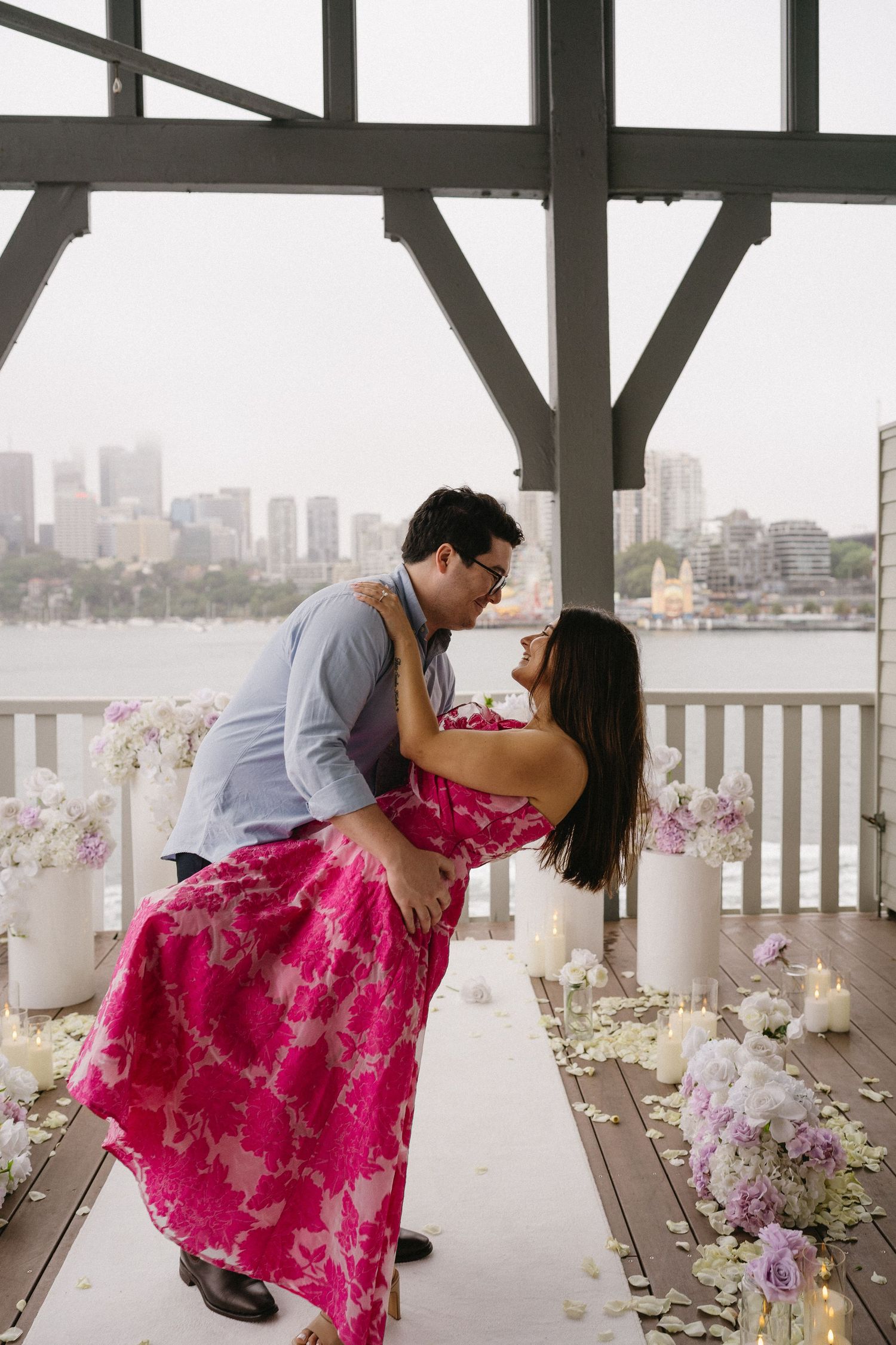 A couple dips romantically surrounded by flowers and candles with a city skyline backdrop.