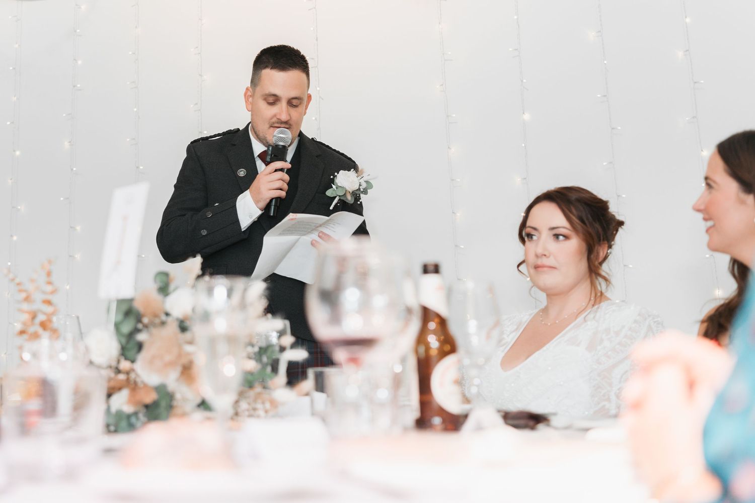 A wedding reception scene with guests seated at a decorated table while someone gives a speech at the celebration.