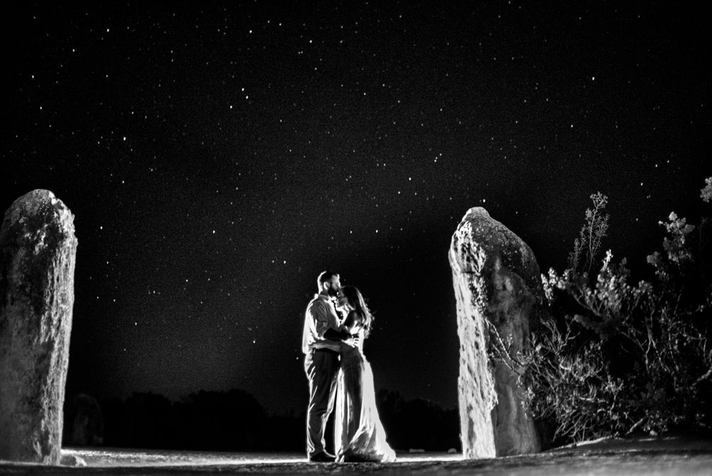 Couple embracing between stone pillars under starry night sky. Lancellin sand dunes