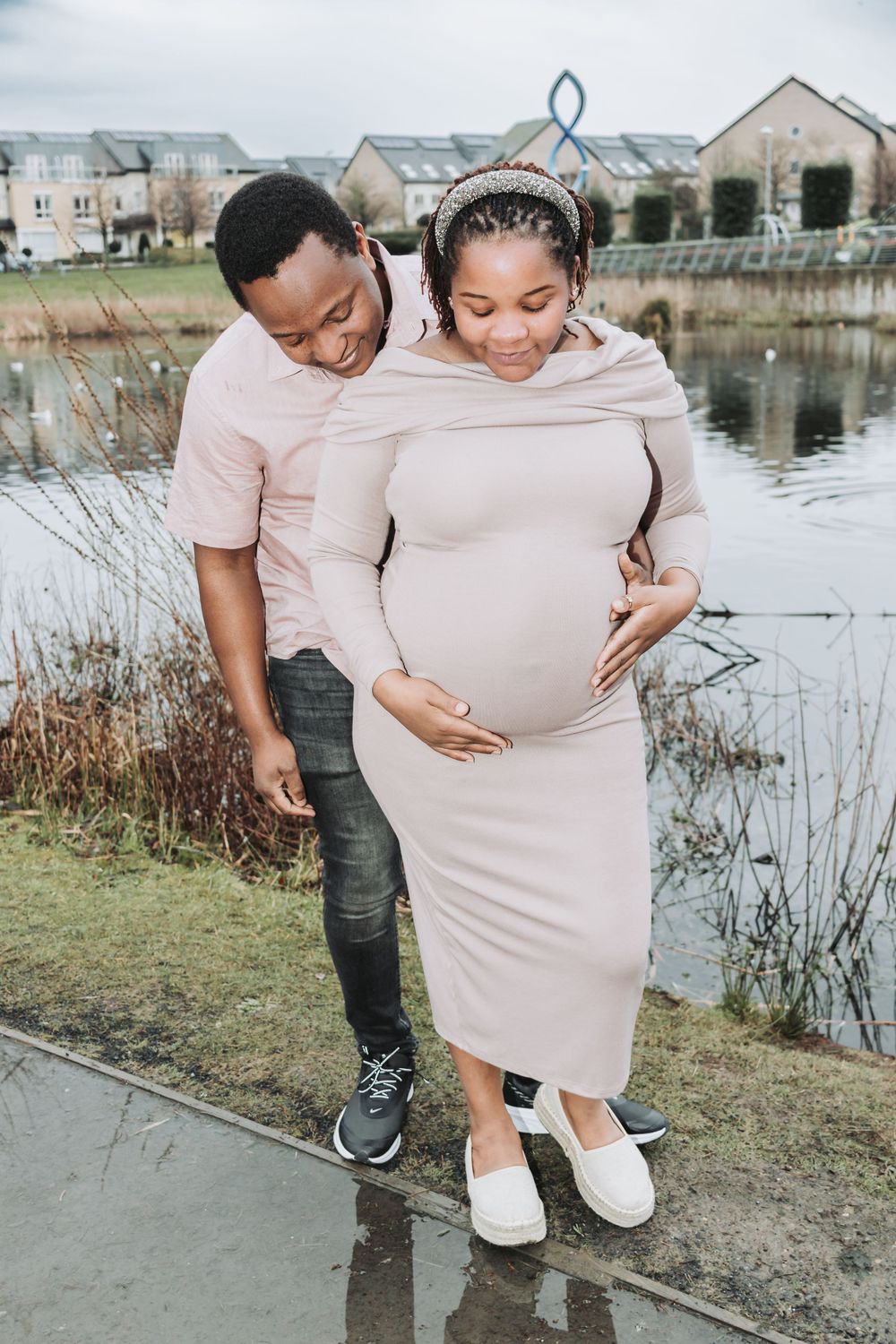 A couple shares an intimate moment by a pond as they celebrate their pregnancy wearing matching neutral-toned outfits.