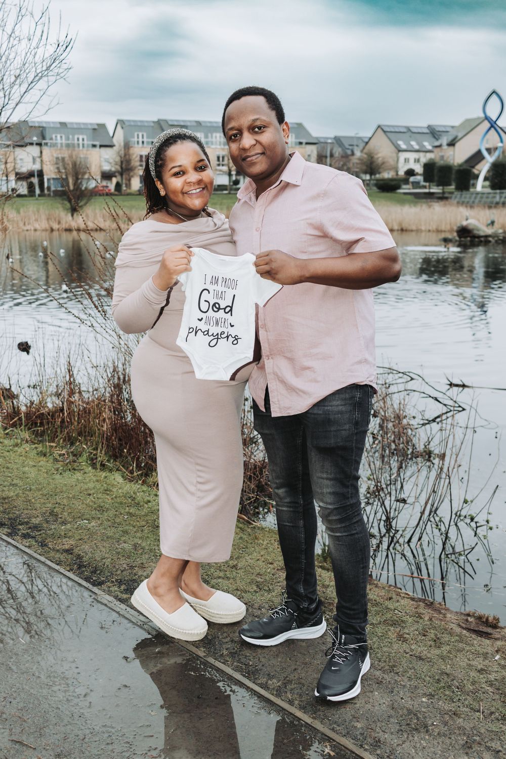 A couple stands by a pond holding a baby onesie with text, wearing coordinating pink and neutral toned outfits.