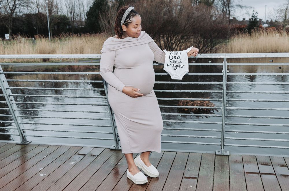 An expectant mother in a light mauve dress holds a tiny white onesie while standing on a wooden dock by the water.