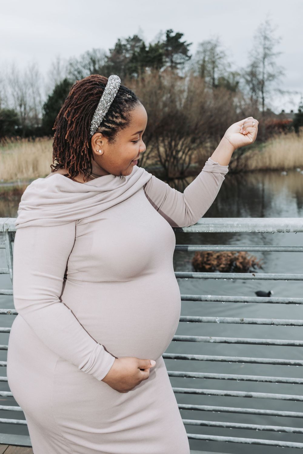 Expecting mother in a light beige dress stands on a wooden dock by a lake during autumn.