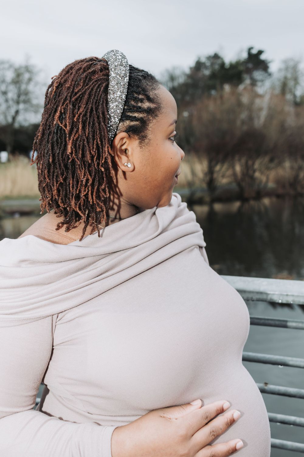 A person wearing a beige sweater and sparkly headband sits on a bench near a pond, photographed from behind.