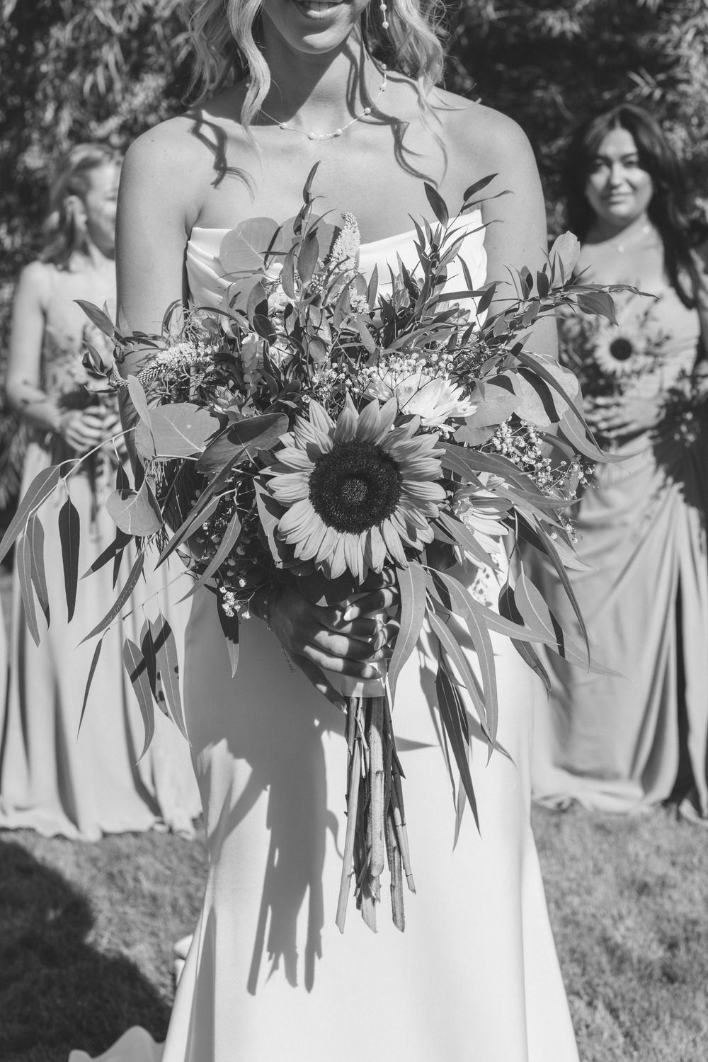 Row of bridesmaids in light dresses holding beautiful sunflower bouquets outdoors.
