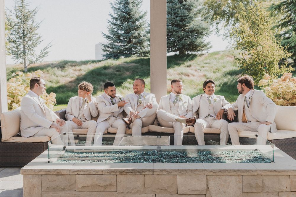 Wedding party members in light beige suits sit casually on a stone wall during an outdoor celebration.