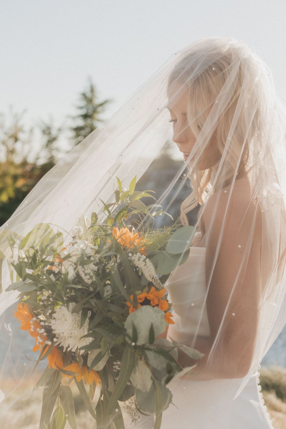 Bride in white veil holding sunflower bouquet against bright outdoor background.