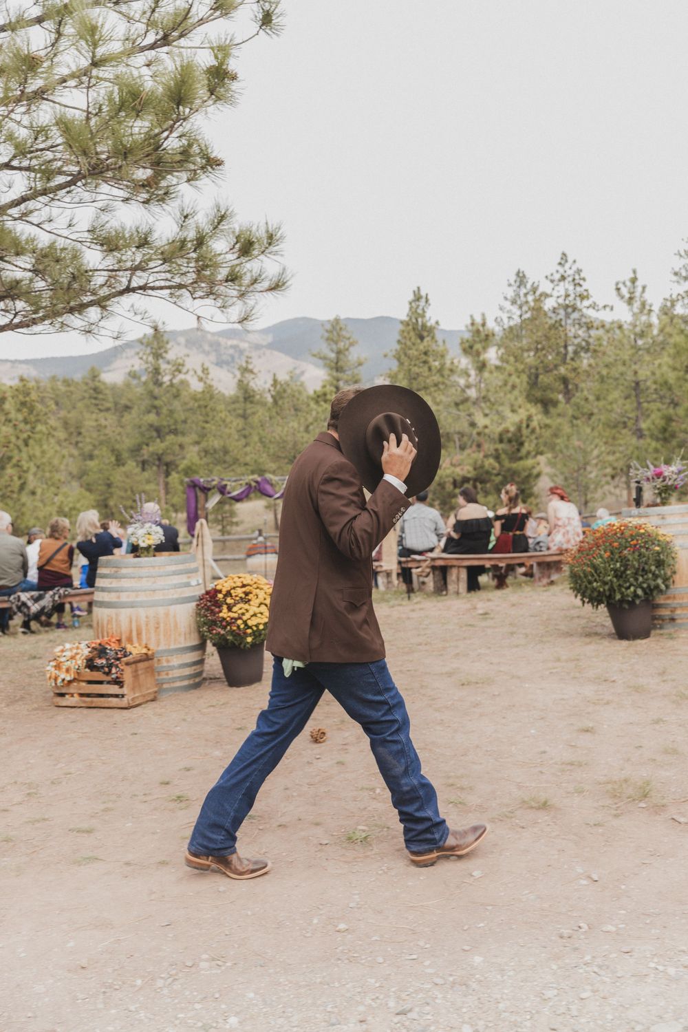 A person in a brown jacket and jeans performs a silly dance sequence outdoors near pine trees and fall decorations.