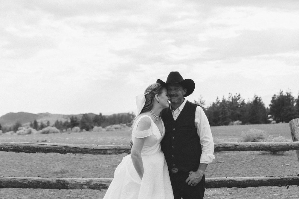 A couple shares an intimate moment in a scenic outdoor setting with mountains and fields in the background.